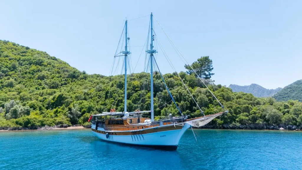 Two-masted wooden sailing yacht anchored in a turquoise bay with forested hills behind it.