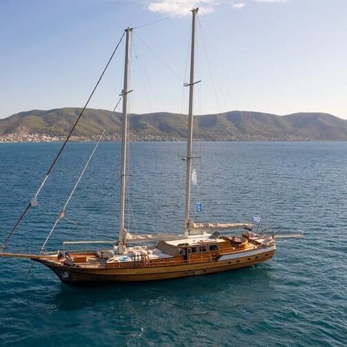 charter-entre-cielos-yacht-2 - Copy | Sail in Greece™ A wooden two-masted sailing yacht moored on calm blue water near a hilly coastline under a clear sky.