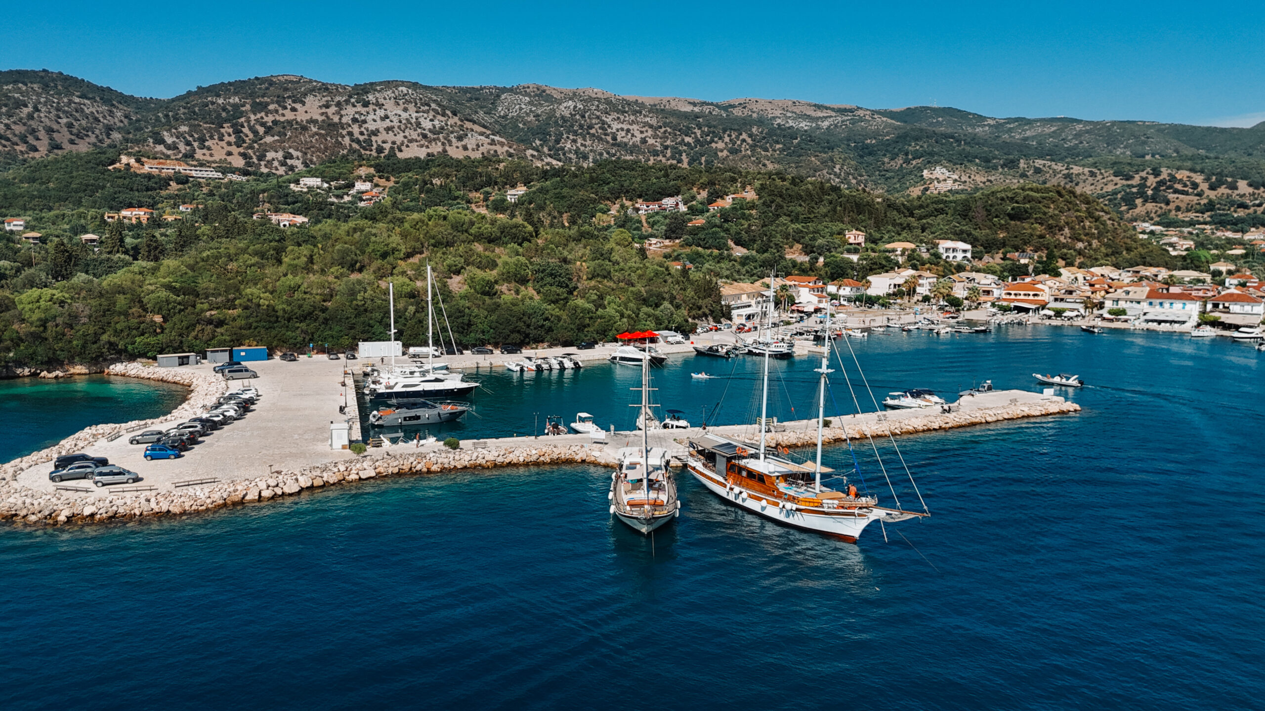 Marina with sailboats and yachts docked along a stone breakwater, hills and a hillside town in the background under a blue sky.