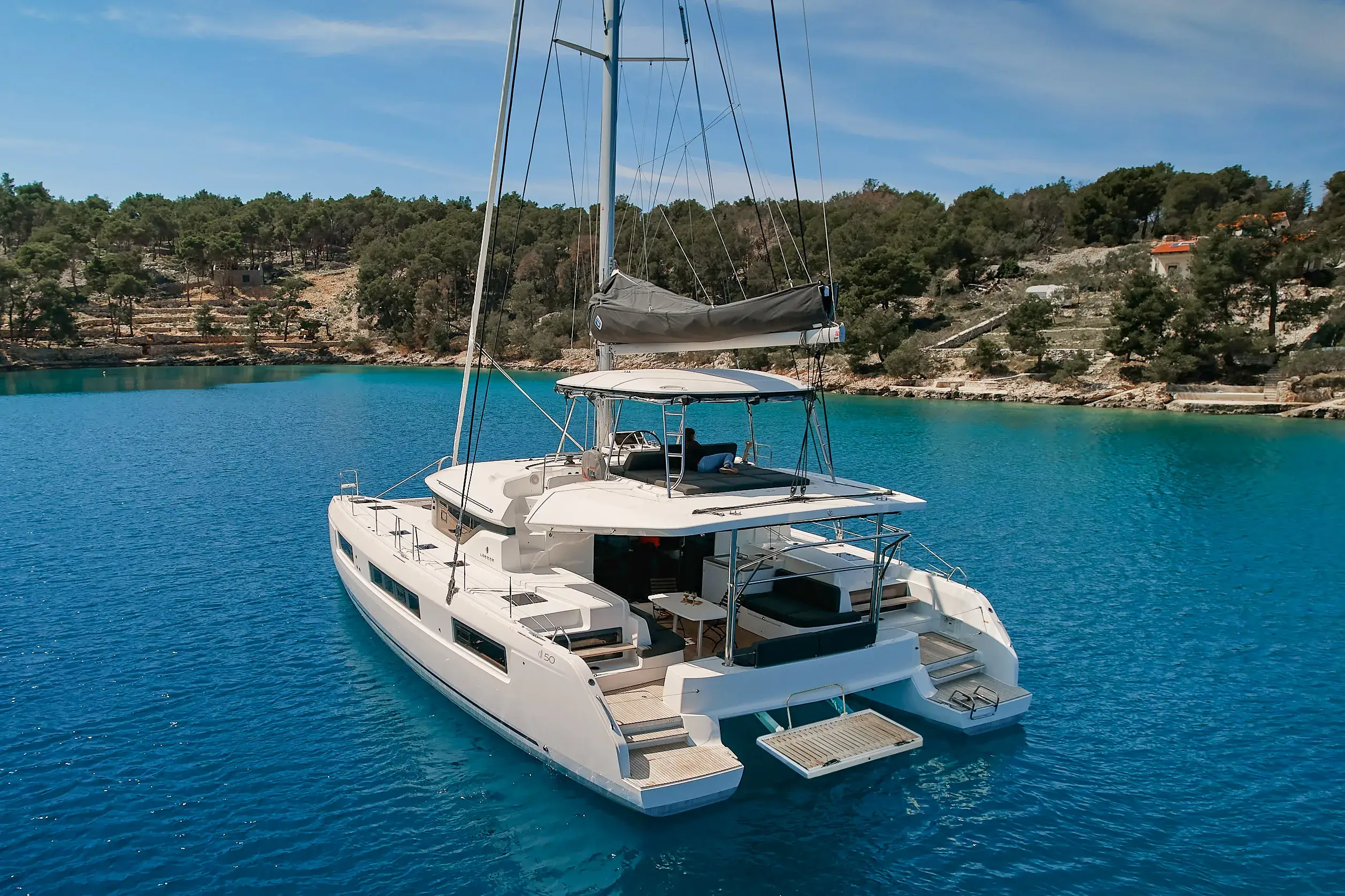 White catamaran on turquoise water with tall masts, rocky shoreline and pine trees in the background.