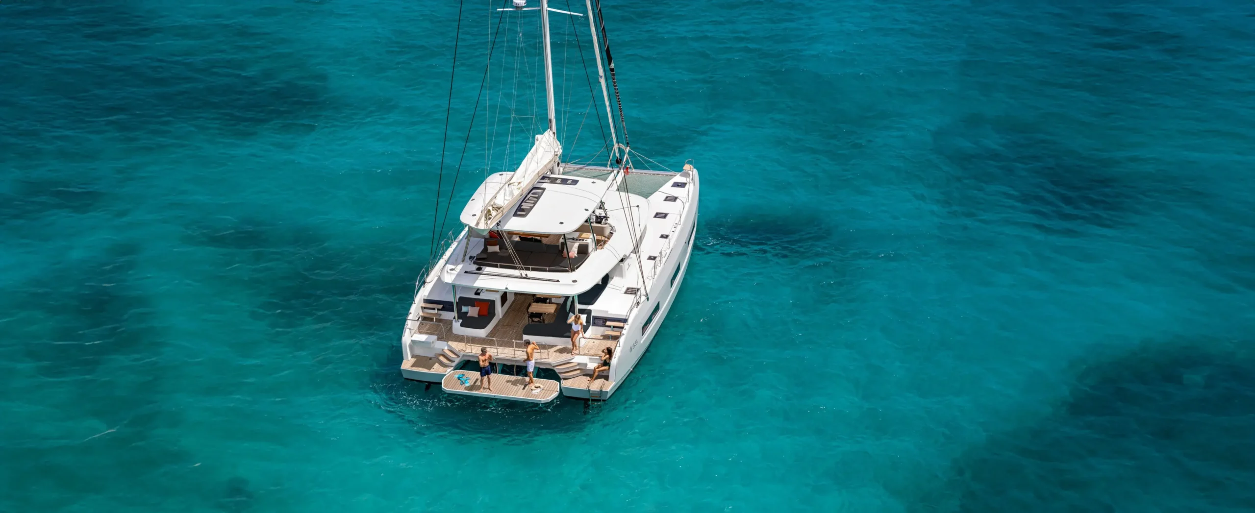 Aerial view of a white catamaran sailing boat with people on the aft deck in clear turquoise water.