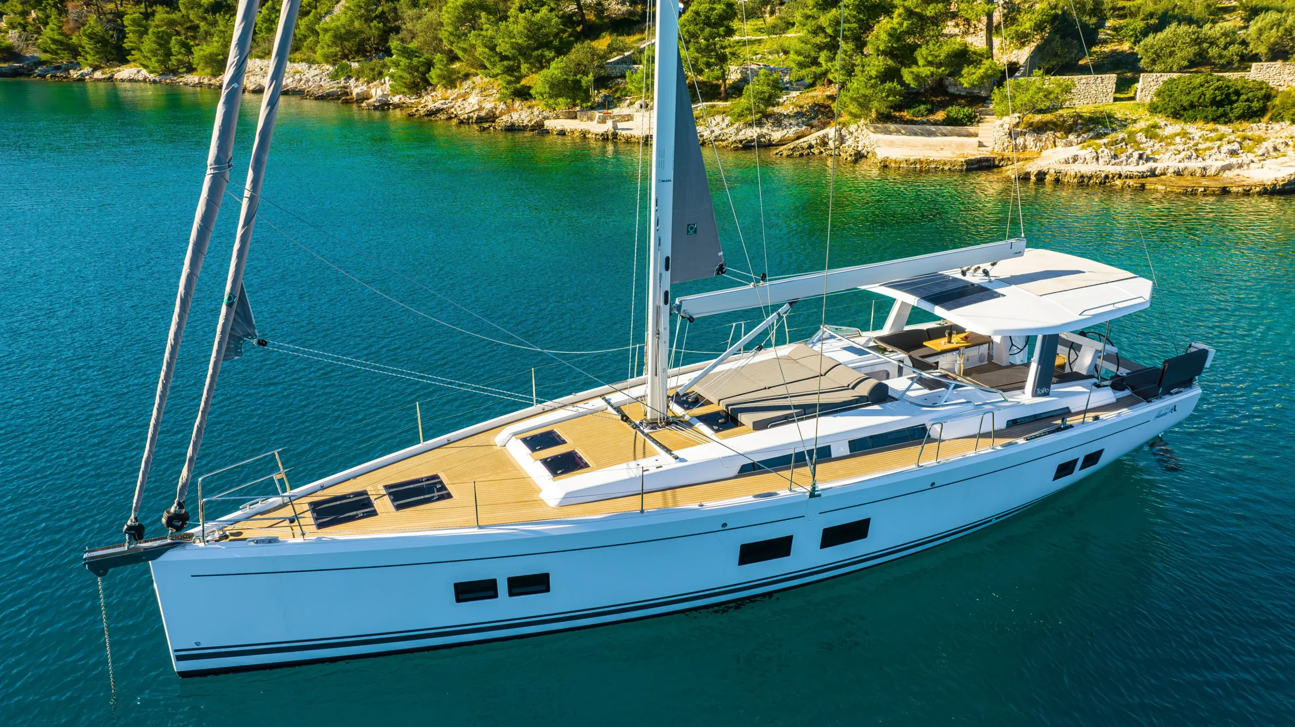 White catamaran anchored in turquoise bay with rocky, tree-covered shore in the background, viewed from above, calm water around it.
