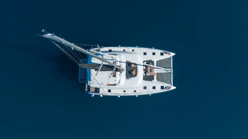 Top-down view of a white sailing catamaran on dark blue water with rigging and two hulls visible, and people on deck beneath a clear sky.