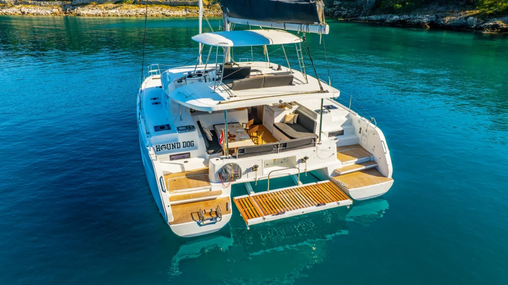 White luxury catamaran yacht with an outdoor dining area on the rear deck, turquoise water around, and a black canopy above; name 'HOUND DOG' visible on the stern.