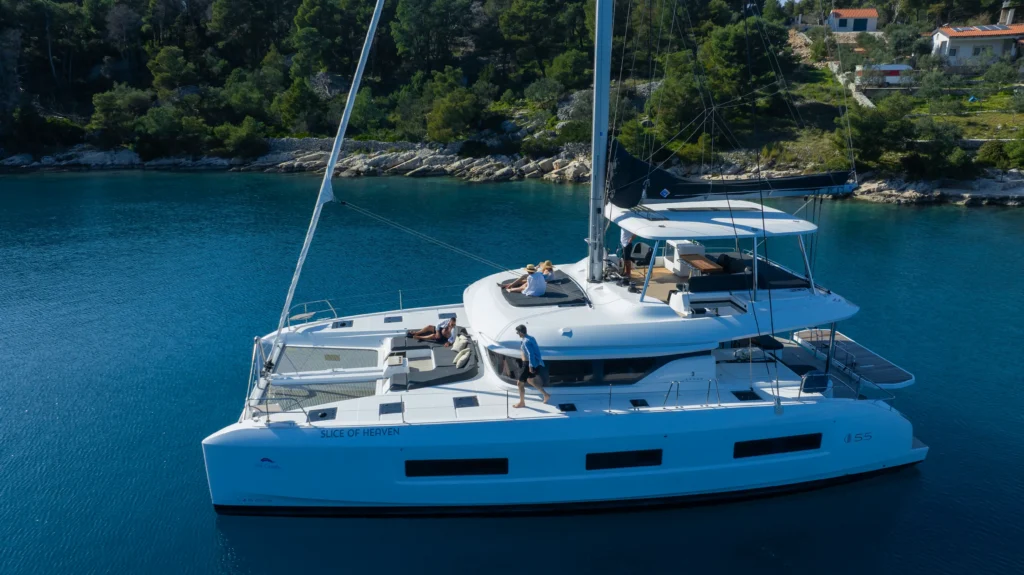 White catamaran on blue water with people lounging on the deck along a rocky coastline.