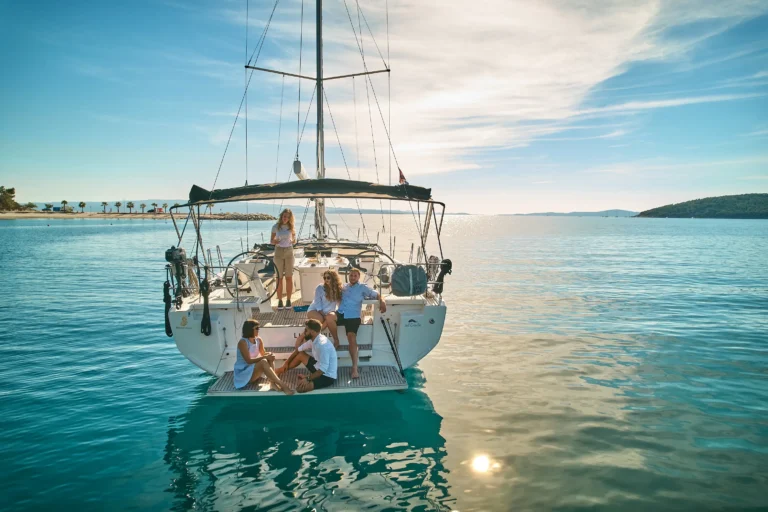 Group of people relaxing on the stern of a sailboat anchored in turquoise water with a coastal shoreline in the background