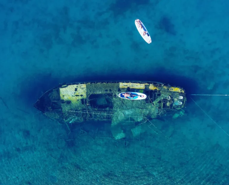 Aerial top-down view of a sunken shipwreck on the seabed, with two stand-up paddleboards floating above in clear blue water.