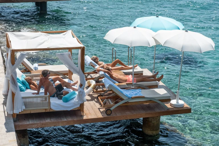 Sunbathers on a wooden floating deck with white loungers and umbrellas over clear turquoise water.