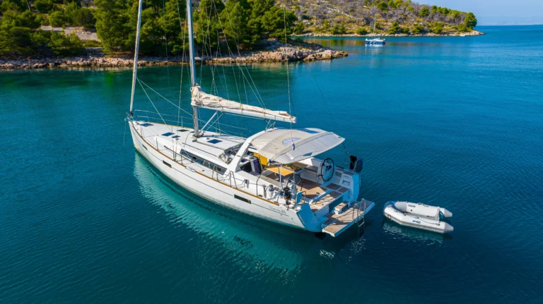 White sailboat anchored in clear turquoise water near a rocky green shoreline, with a small inflatable dinghy moored alongside.