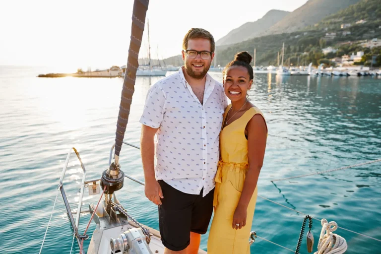 Smiling couple standing on a sailboat at sunset by a marina, man in a white patterned shirt and woman in a yellow dress.
