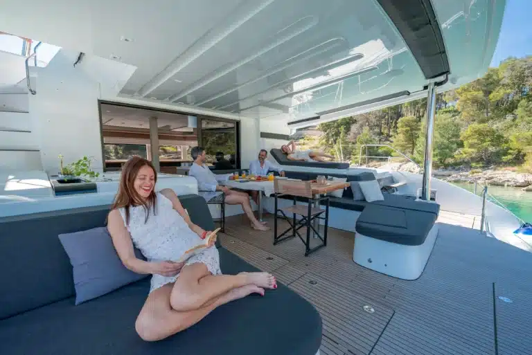 People relaxing on a luxury yacht deck with cushioned seating, a table set for a meal, and friends chatting on a sunny day by the water.
