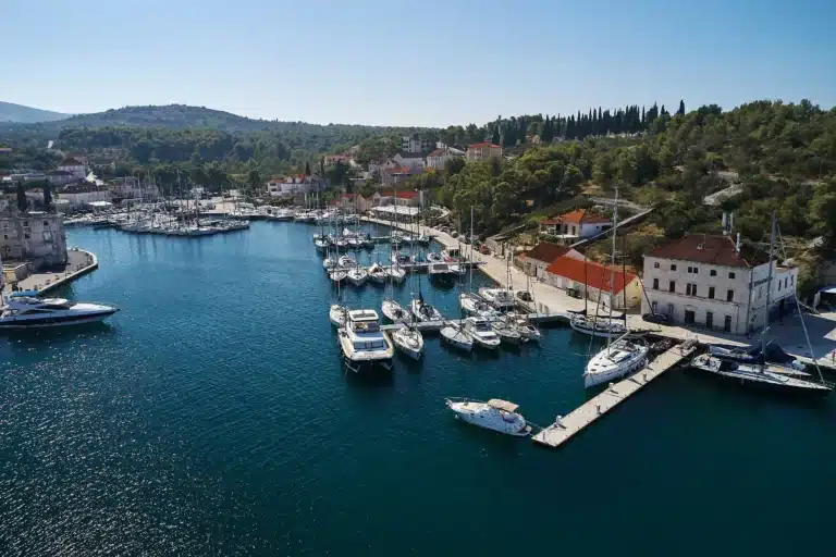 Harbor with many moored sailboats and yachts, red-roofed buildings along the shore, and forested hills in the background.