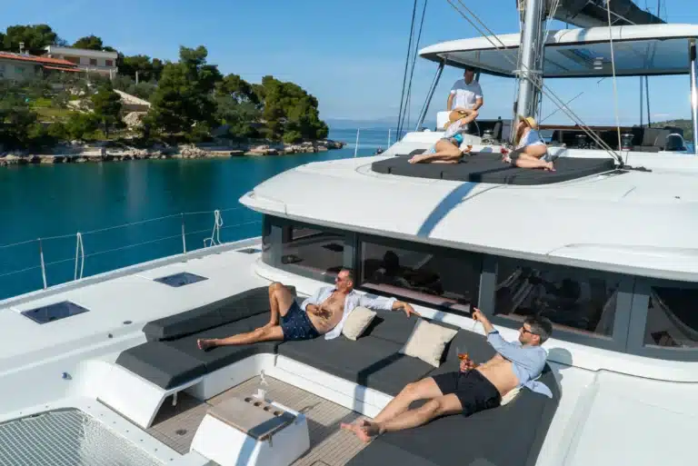 People lounging and sunbathing on the curved sun deck of a white luxury yacht, blue sea and a rocky, tree-covered coastline in the background.