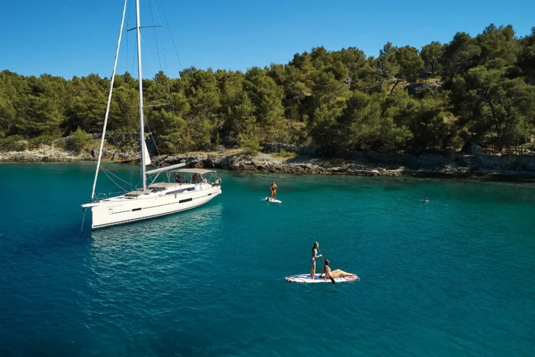 White sailboat anchored in turquoise water beside a rocky, forested coastline; people paddleboarding nearby on a sunny day.
