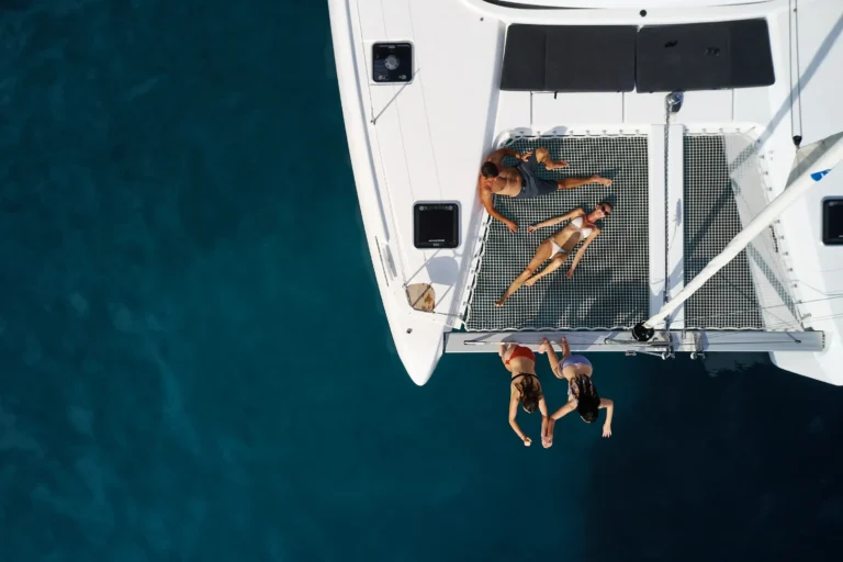 Top-down view of people sunbathing on the netting of a white catamaran above deep blue water