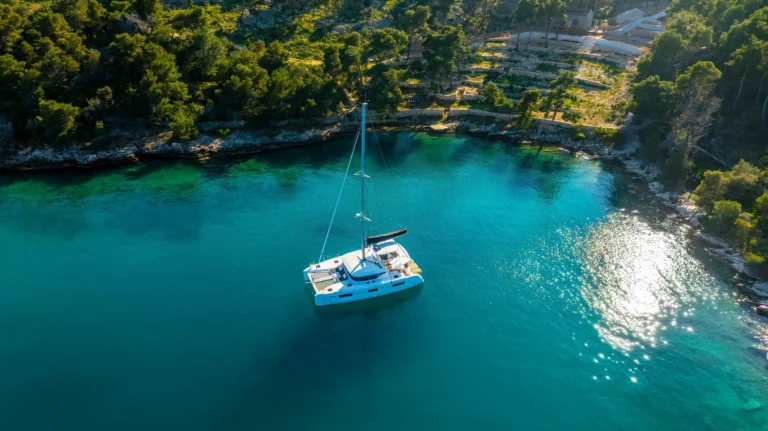 A white sailboat on turquoise water near a rocky, tree-lined coastline (aerial view).
