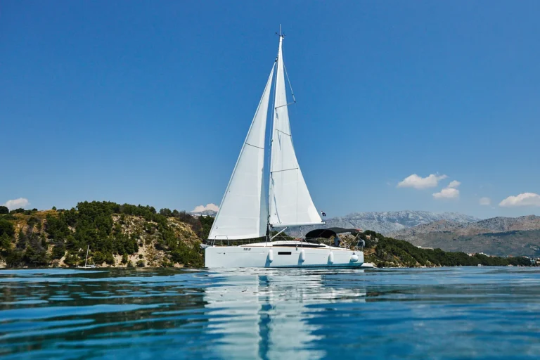 White catamaran sailing on calm blue water with a rocky green coastline and distant mountains under a clear sky.