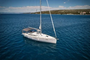 White catamaran anchored in calm blue sea with a rocky, tree-lined coastline and hills in the distance under a clear sky.