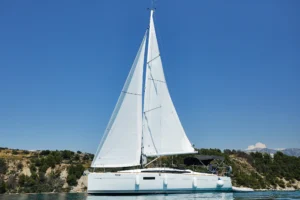 gallery-Manilo-3 | Sail in Greece™ White sailing catamaran with tall sails on calm blue water near a rocky coastline under a clear sky.