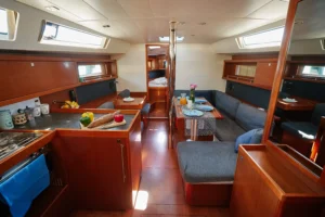 Sailboat cabin with polished wood cabinetry, a dining table set for meals, and a galley area with fruit on the counter on the left. The space leads to a sleeping area in back via a doorway.