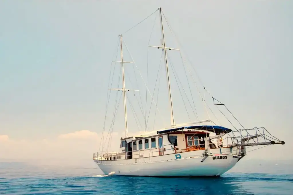 White two-masted sailboat on calm blue water, name 'Glaros' on the bow with a blue canopy over the cockpit.