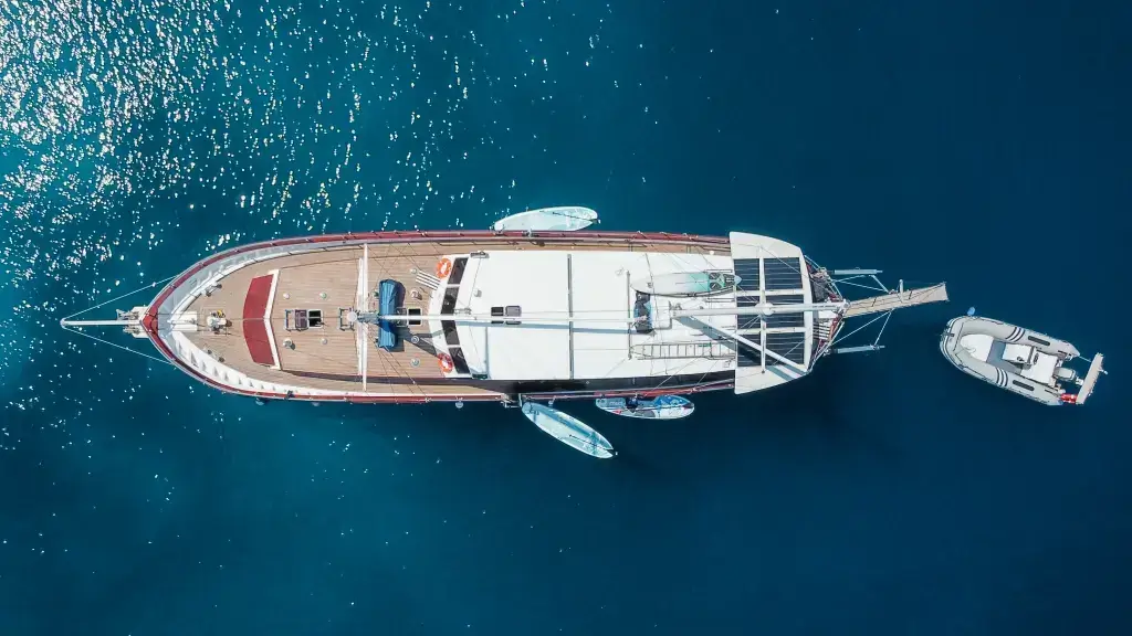 Overhead view of a long wooden luxury yacht anchored in blue water, with a small dinghy nearby on the starboard side.