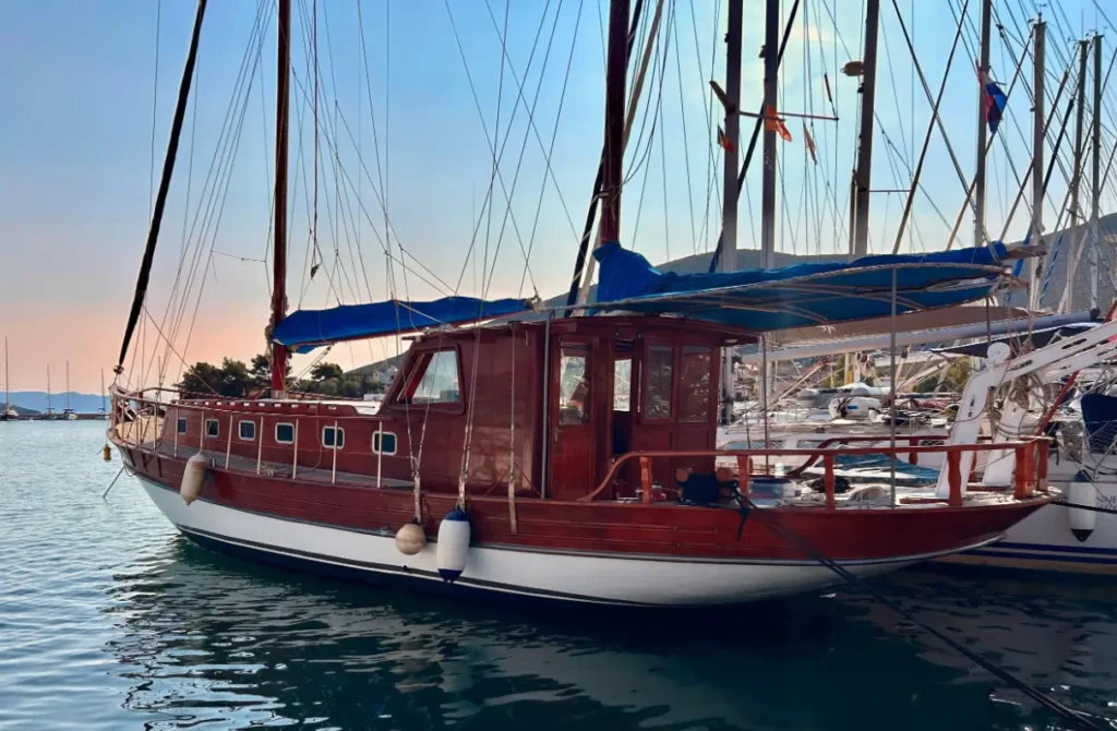 Wooden sailboat with a red-brown hull docked in a marina, blue canopy over deck and tall masts with rigging.
