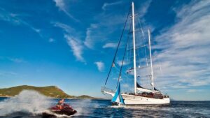 A white two-masted sailing yacht on blue water, with a jet ski spraying water nearby and green hills in the distance under a clear sky.