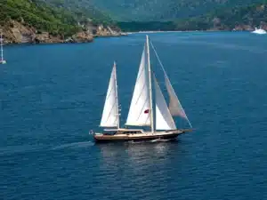 Two-masted sailboat with white sails gliding across blue sea near a rocky, forested coastline.