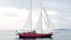 Red two-masted sailboat gliding on calm water with people on deck and a Greek flag at the stern.