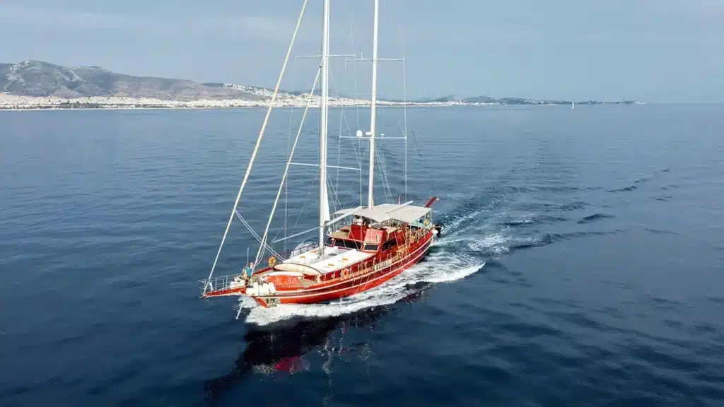 Red sailboat cruising on calm blue sea with distant coastline and hills in the background, mast towering above.