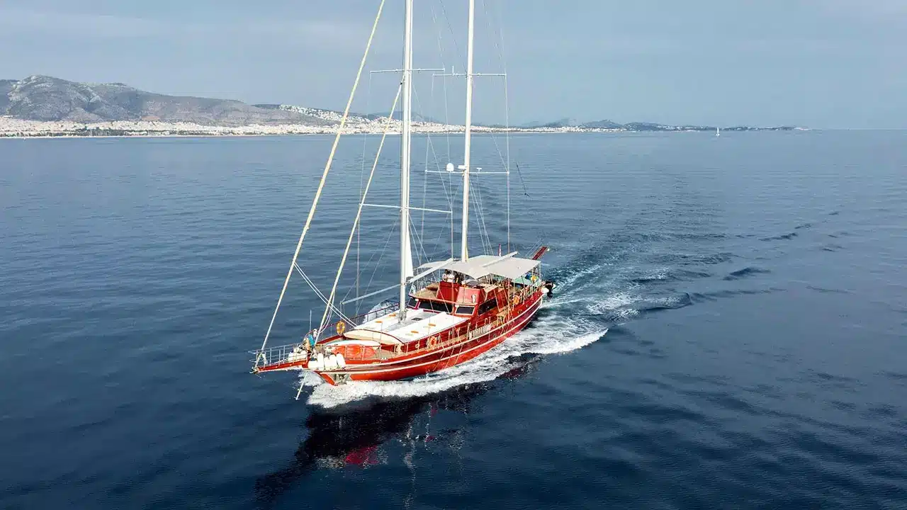 Red sailboat cruising on calm blue sea with distant coastline and hills in the background, mast towering above.