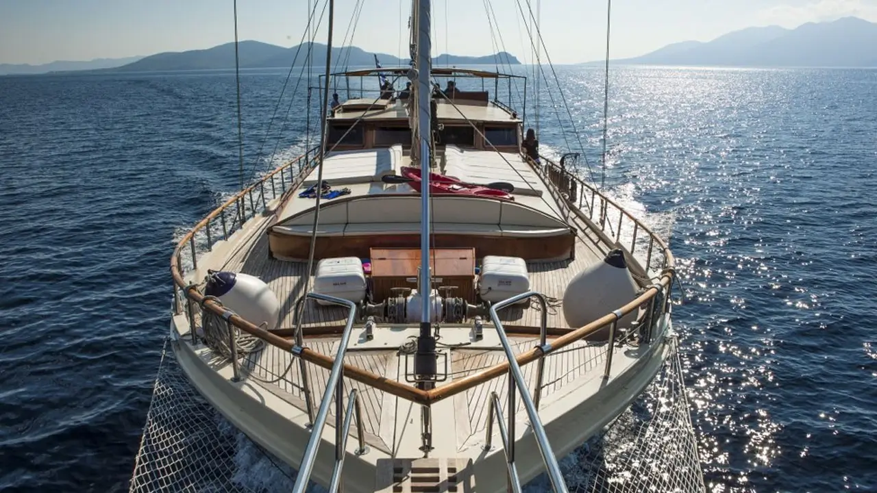 Bow view of a sailing yacht at sea with a wooden deck, rigging overhead and calm blue water extending to distant mountains.