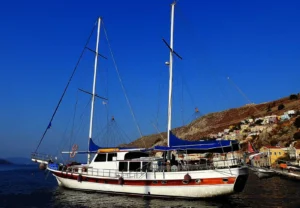 A white and brown sailing yacht moored in calm harbor with a hillside town in the background and a clear blue sky.