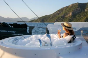 Woman in a straw hat lounging in a bubbling tub on a yacht, holding a glass of wine, with sea and hills behind.
