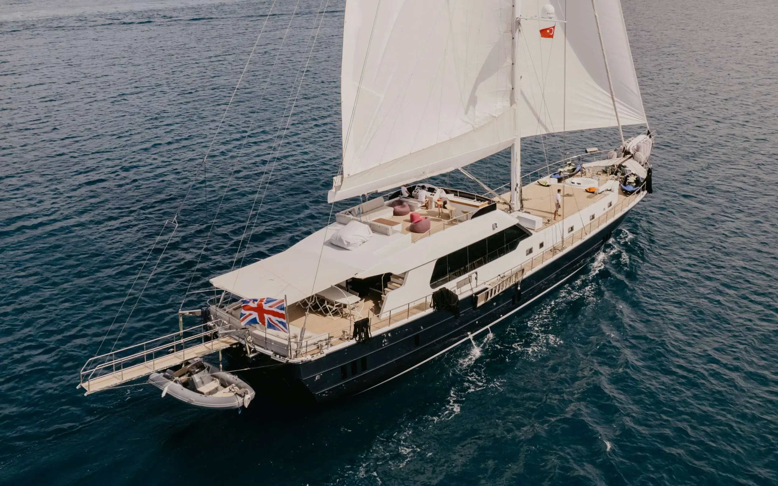 A white sailing yacht with sails up cruising on deep blue water, crew on deck near the cockpit, British flag at the stern.