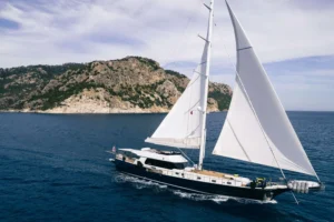 Sailboat with white sails gliding near a rocky island and calm blue sea under a clear sky