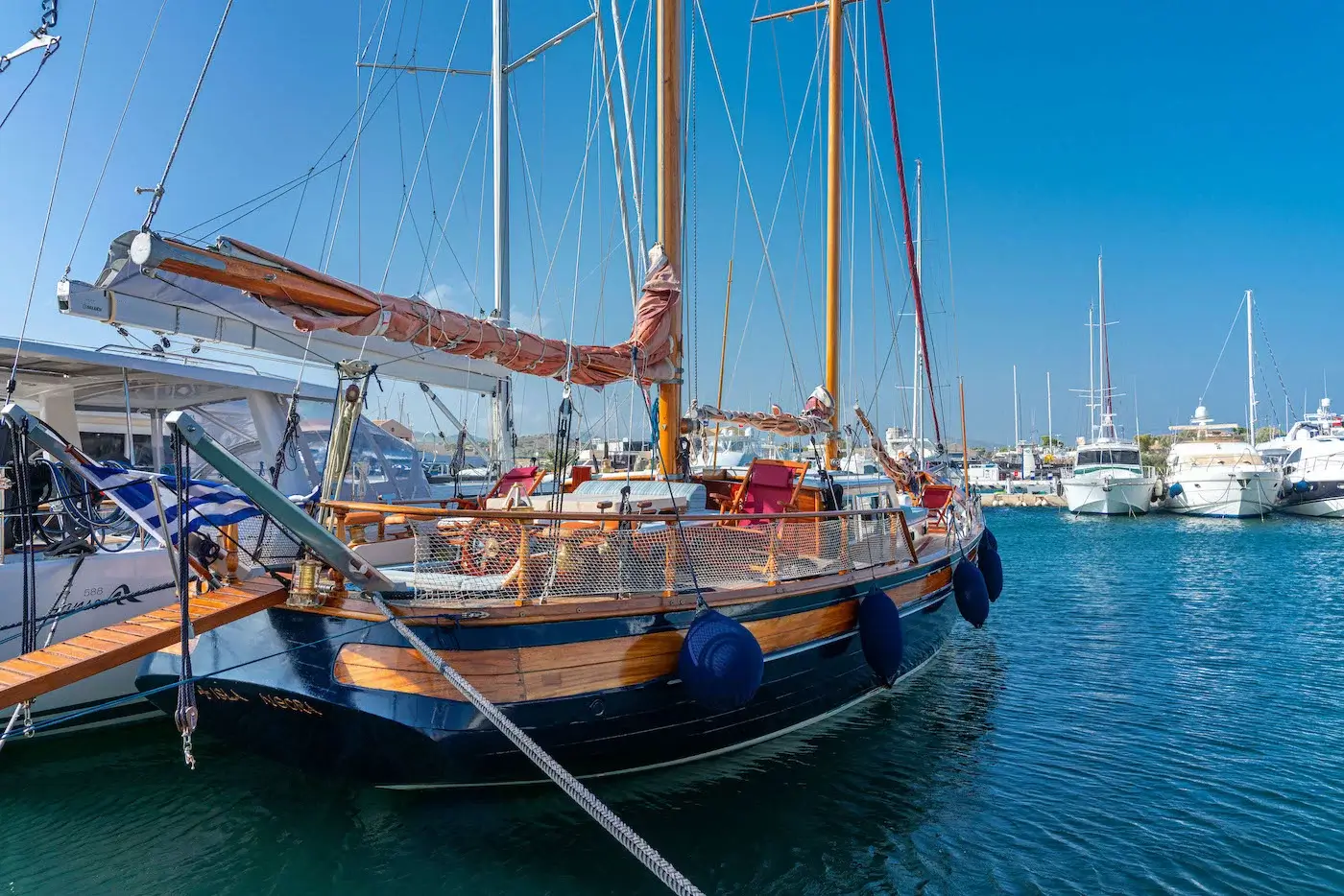 Sailboat with wooden trim docked at a marina, blue water and other yachts in the background under a clear sky.