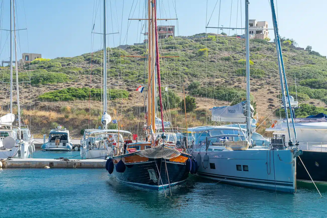 Marina with several sailboats docked side by side and calm blue water in the foreground, hillside with sparse vegetation in the background.