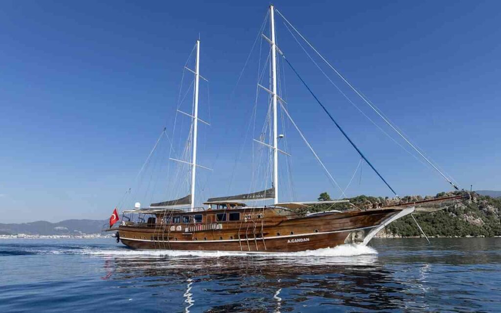 Wooden two-masted sailing yacht with a Turkish flag gliding on calm sea near a rocky shore.