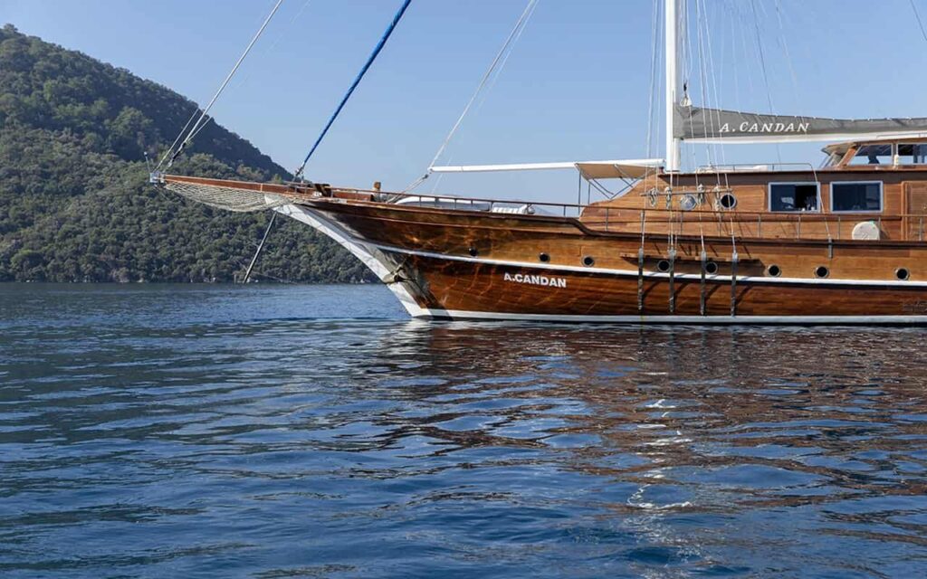 Wooden sailing yacht on calm blue water with forested hills in the background; hull reads 'A. Candan'.