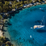 product_yacht_charter_bb63ac77a4 | Sail in Greece™ Aerial view of a turquoise bay with a white sailboat anchored near a rocky, pine-covered shoreline.