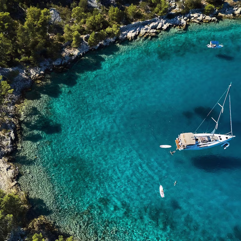Aerial view of a turquoise bay with a white sailboat anchored near a rocky, pine-covered shoreline.