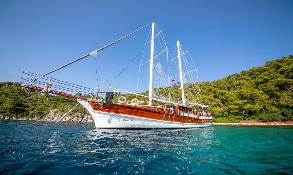 Two-masted wooden sailing yacht anchored near a forested coastline on turquoise water under a blue sky.