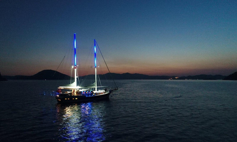 Sailboat with blue-lit masts on calm sea at dusk, reflecting lights on the water over distant hills on the horizon.