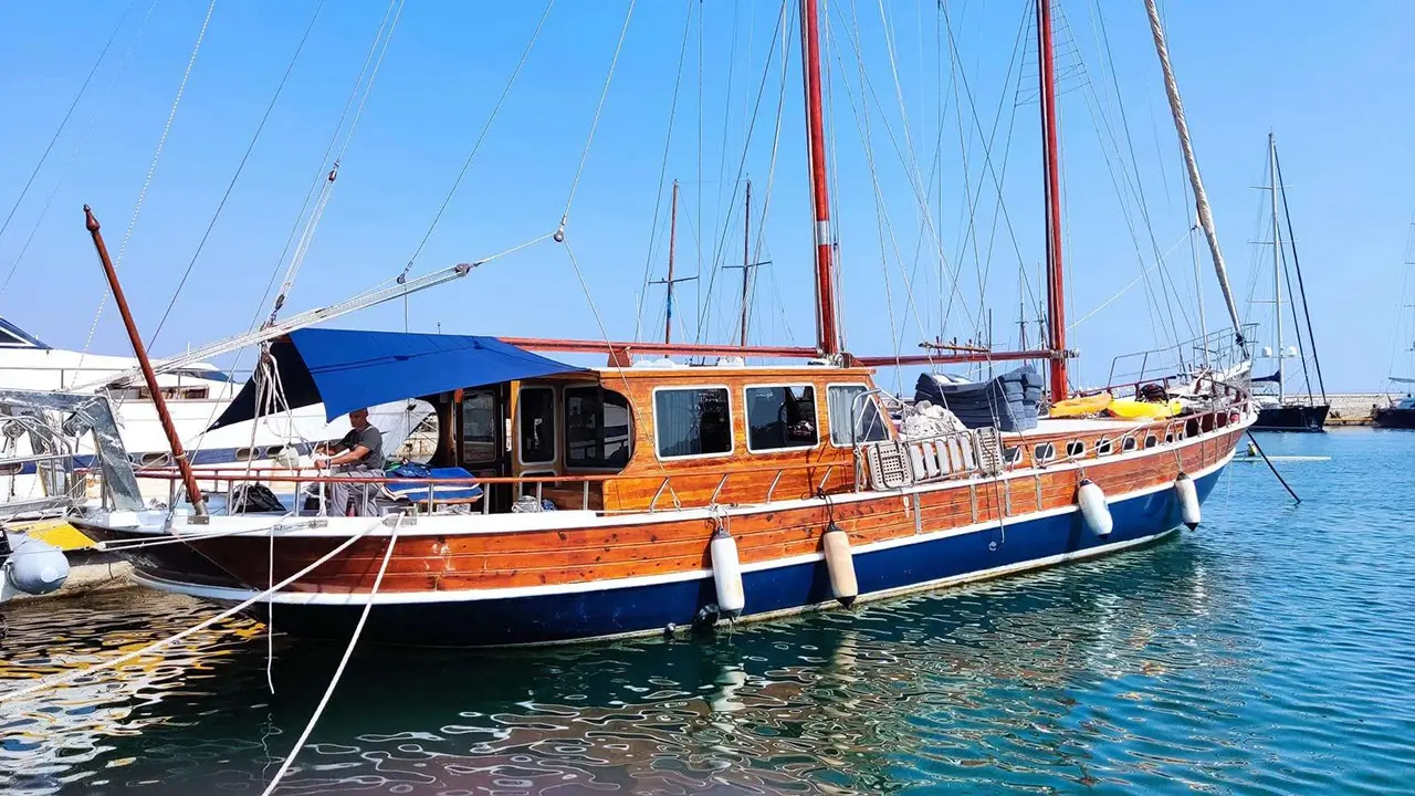 Wooden sailing yacht moored at a marina under a blue sky, with a blue canopy over part of the deck and masts looming above.