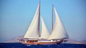 stella-1 | Sail in Greece™ Twin-masted wooden sailboat with white sails gliding on blue ocean, distant hilly coastline in the background”,