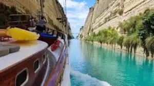 A wooden boat cruises through a narrow turquoise canal between high rocky cliffs and palm trees on the right.