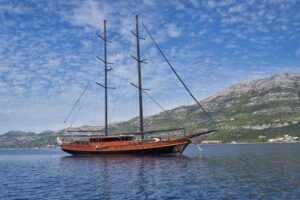 Two-masted wooden sailing ship on calm blue water with mountains in the distance in the background.