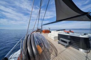 Sailboat deck with from-left lifebuoys, wooden planks, and a shaded cockpit area overlooking calm blue sea on a clear day.